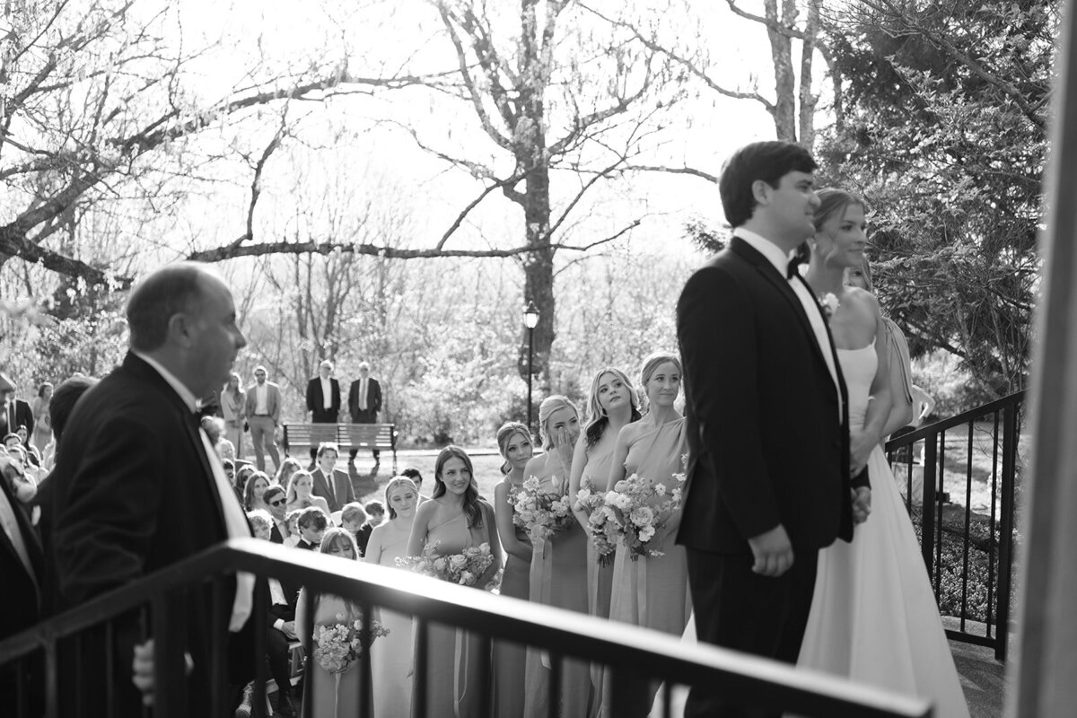 Black and white photo of the bride and groom standing at the altar with the bridal party and guests in the background during the wedding ceremony at Burritt on the Mountain in Huntsville, Alabama, photographed by Perry Hancock.