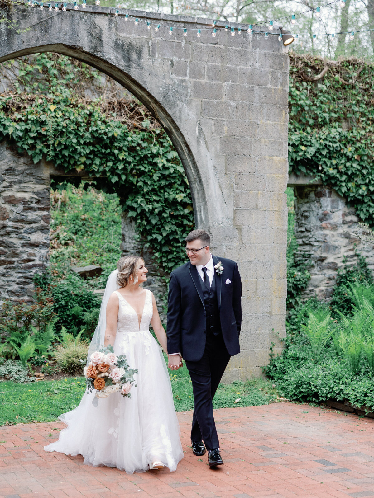 A bride and groom walks together as they gaze into each other's eyes under a stone archway