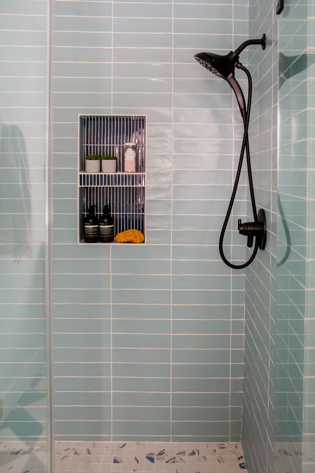A modern shower area tiled with horizontal rows of light blue rectangular tiles and white grout. A matte black showerhead on a curved arm, along with a matching control valve, is mounted on the tiled wall. A built-in niche with dark blue vertical tiles on the back wall and light blue horizontal tiles on the sides and shelves holds two small potted plants, a light pink bar of soap, two dark bottles, and a yellow sea sponge. The shower floor is tiled with small, irregularly shaped tiles in shades of white and blue. A clear glass shower door is partially visible on the left. 