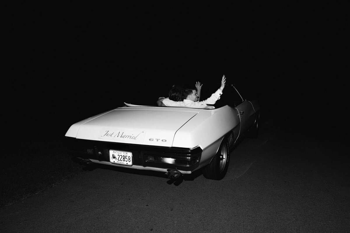 A black and white wedding photo of the bride and groom in their getaway car driving away at the end of their wedding night