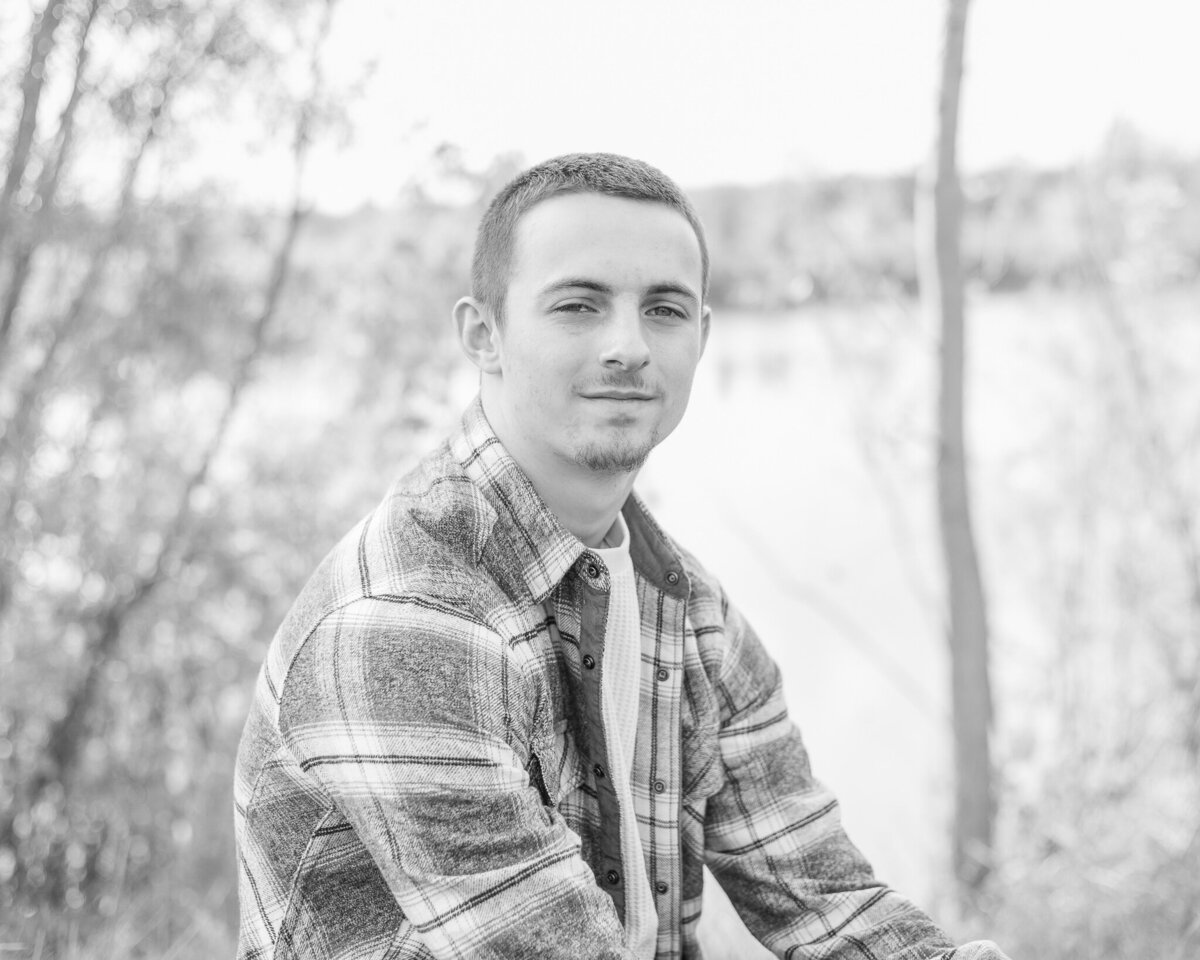 black and white photo of a high school senior male, sitting to the side and in the grass, wearing a flannel jacket , looking at the camera with a serious look