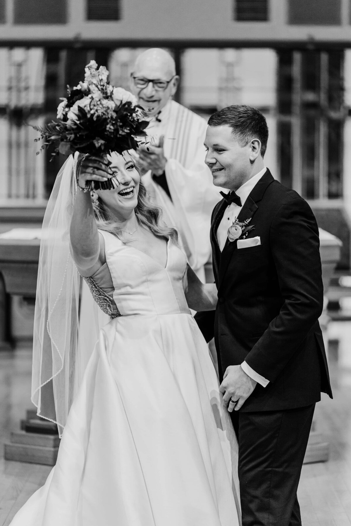 Bride joyfully throws her bouquet, laughing, next to the smiling groom during a wedding ceremony in a park, with a priest observing in the background.