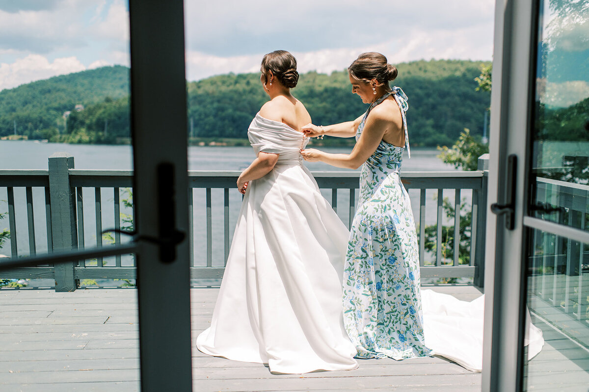 Bride getting zipped into her gown on a deck overlooking Lake Glenville with a bridesmaid in a blue-and-green floral dress.
