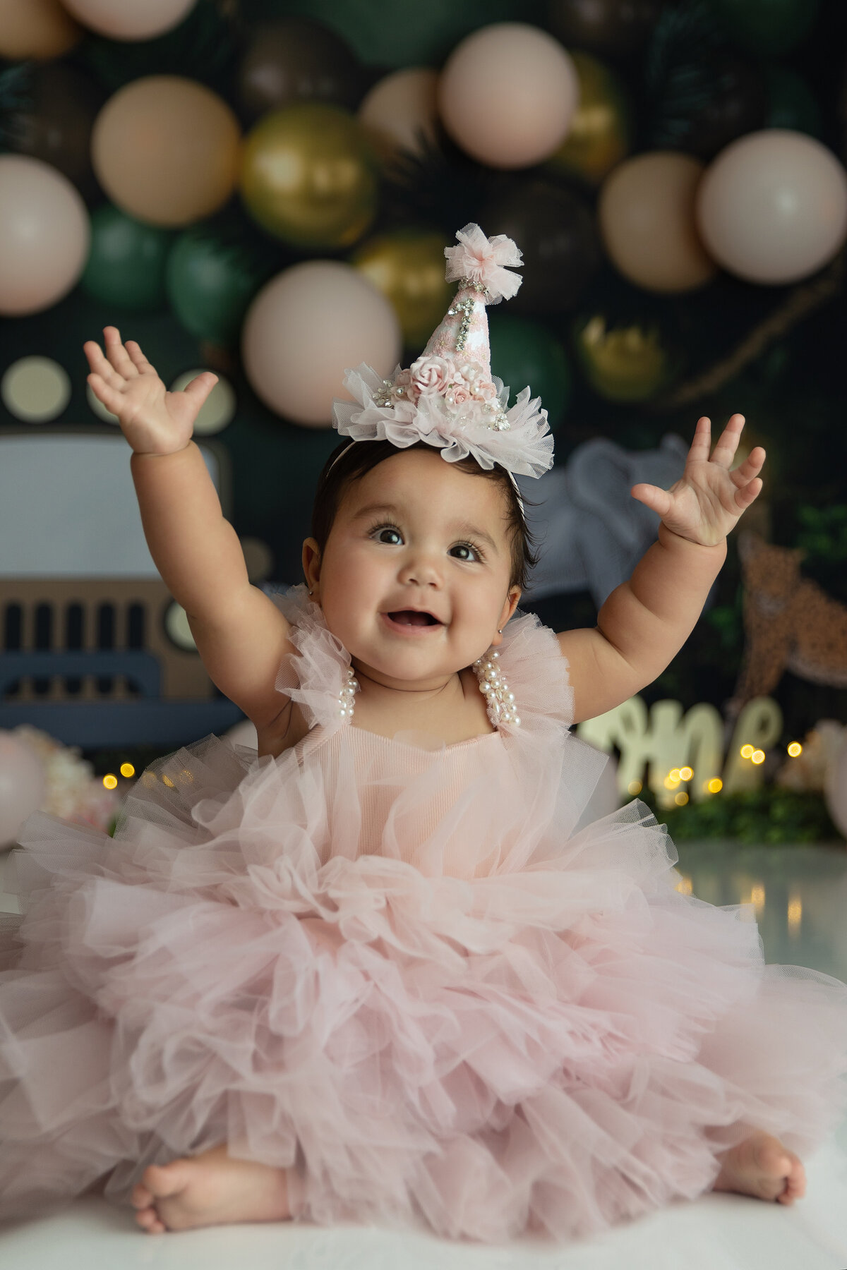 A happy baby girl throws her hands in the air while wearing a pink dress and matching birthday hat