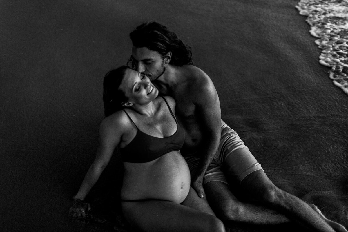 Black and white image of happy expecting couple laying on the beach during their Hawaii babymoon photography session