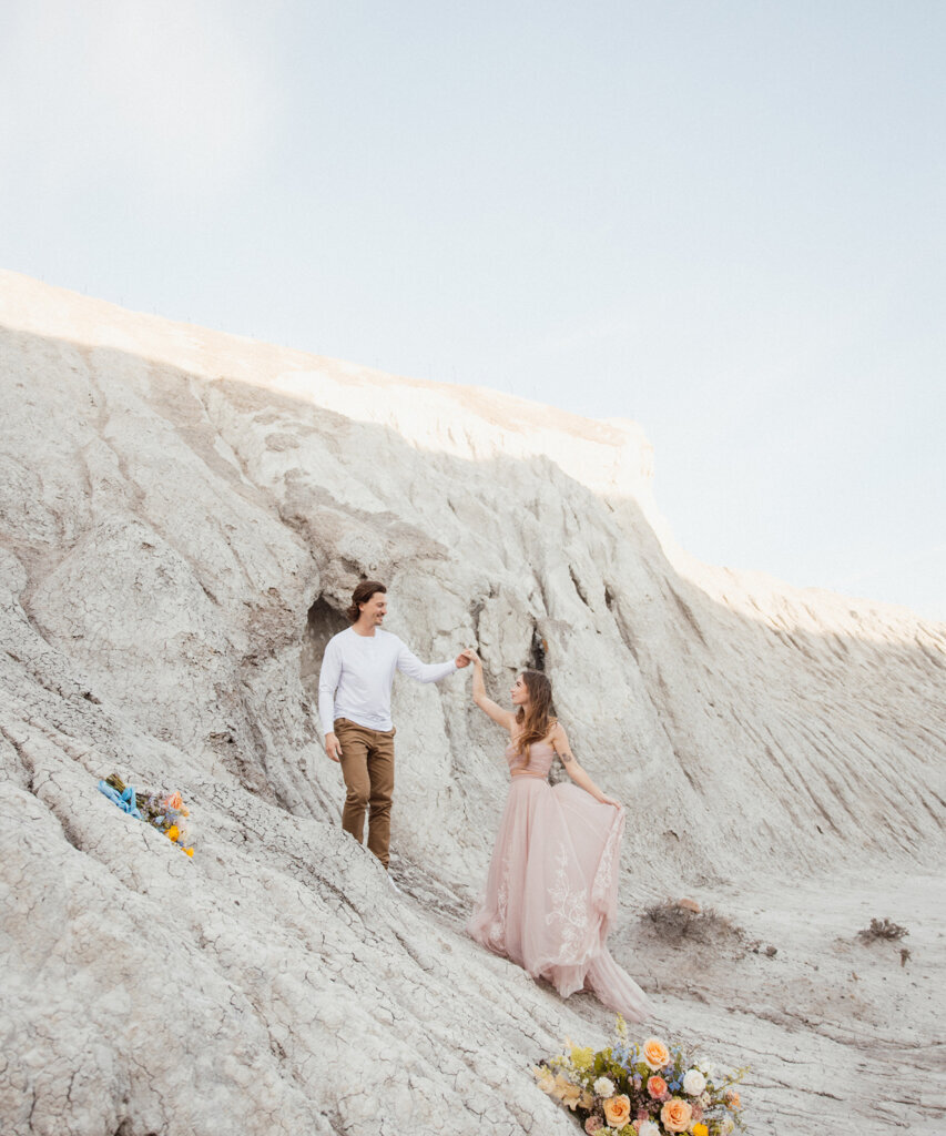 Couple holding hands on a stone hill, woman wearing elegant blush gown captured by Ninth Avenue Studios, authentic and intimate wedding photographer in Calgary, Saskatoon, and Vancouver. Featured on the Bronte Bride Vendor Guide.