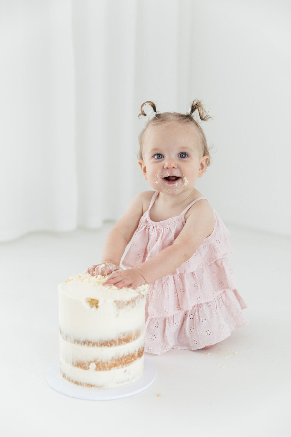 One year old girl in pink dress leans against her birthday cake looks at the camera with a smile
