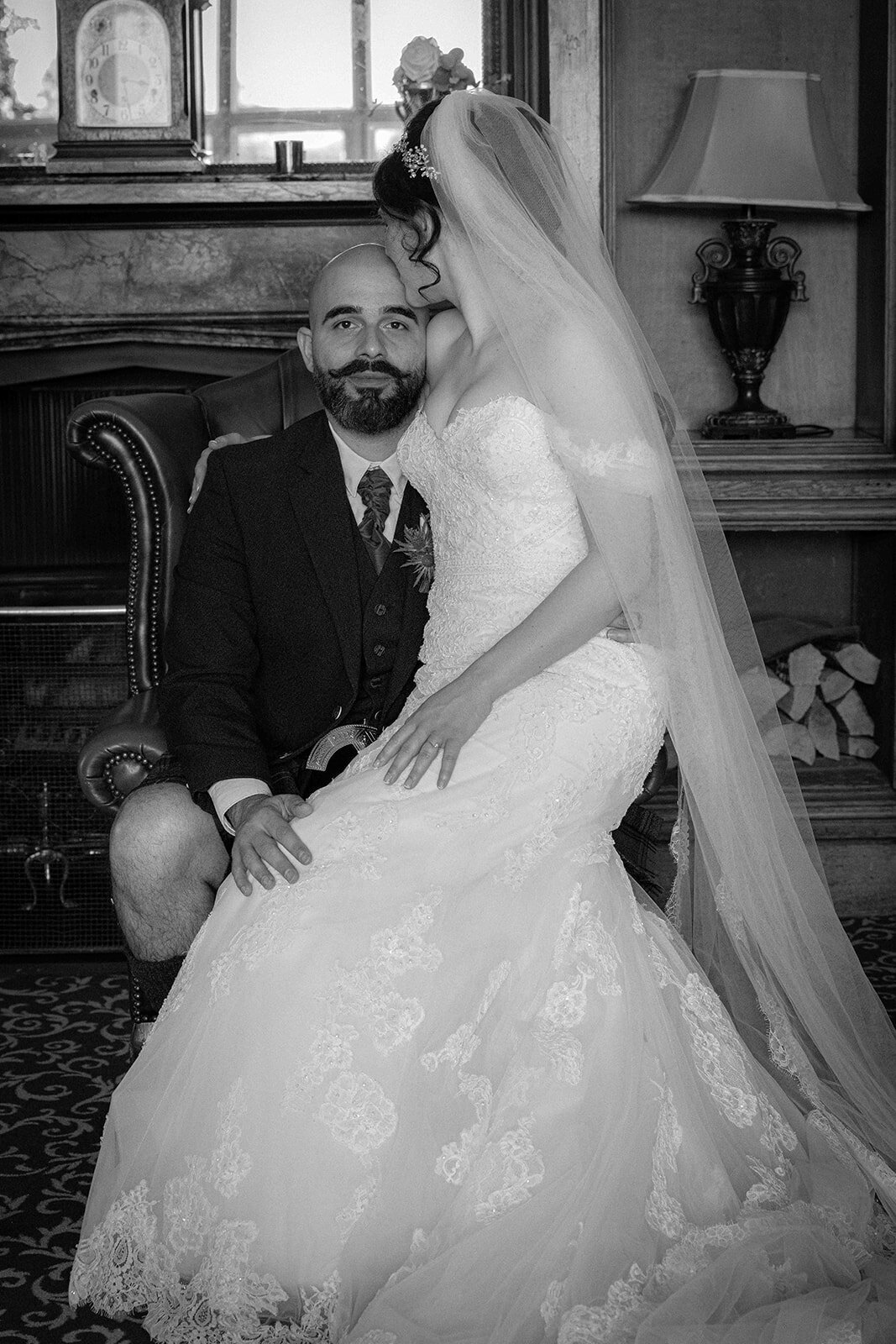 A groom sits looking at the camera with his bride on his knee as she kisses his head.