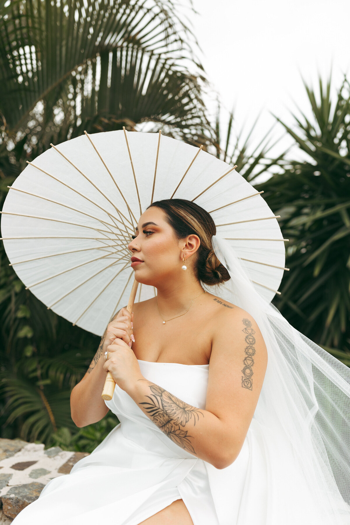 Bride with a parasol looking over her shoulder, her tattooed arm and the details of her white gown highlighted against the greenery