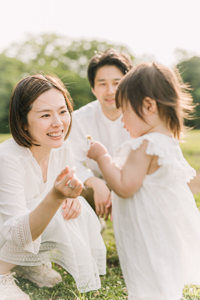 A toddler girl picking up her white clover and her mother and father interacting the girl. Beautiful and light and airy family photograph, shot by photographer Yukika Ishikawa Photography.