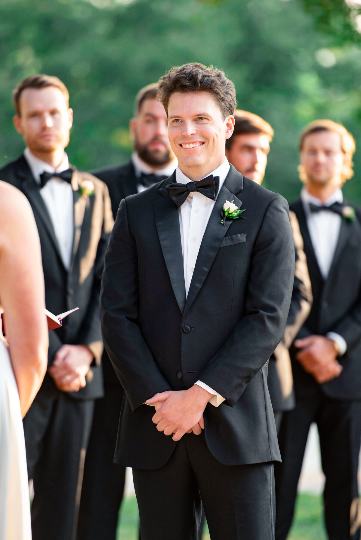A groom in a black tuxedo smiles at the bride during an outdoor wedding ceremony. Five groomsmen stand in the background, wearing matching tuxedos. The setting is lush and green, suggesting a park or garden venue.