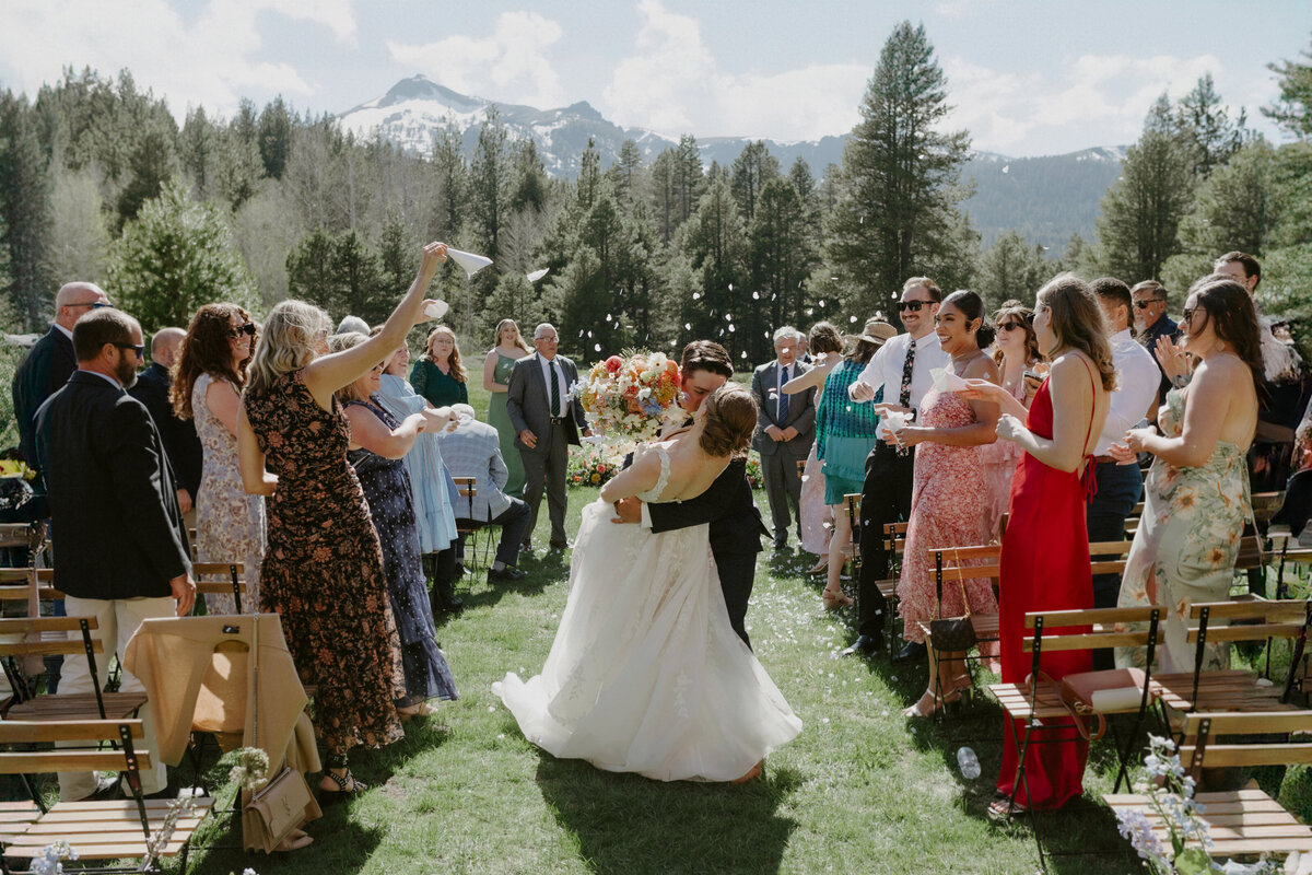 bride and groom walking down aisle in confetti kissing with mountains in background