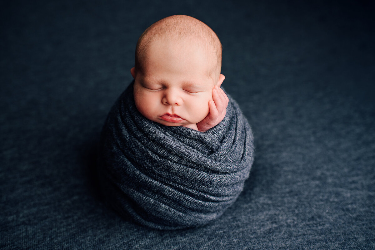Newborn boy wrapped in blue potato pose and hand on cheek in Jacksonville, FL.
