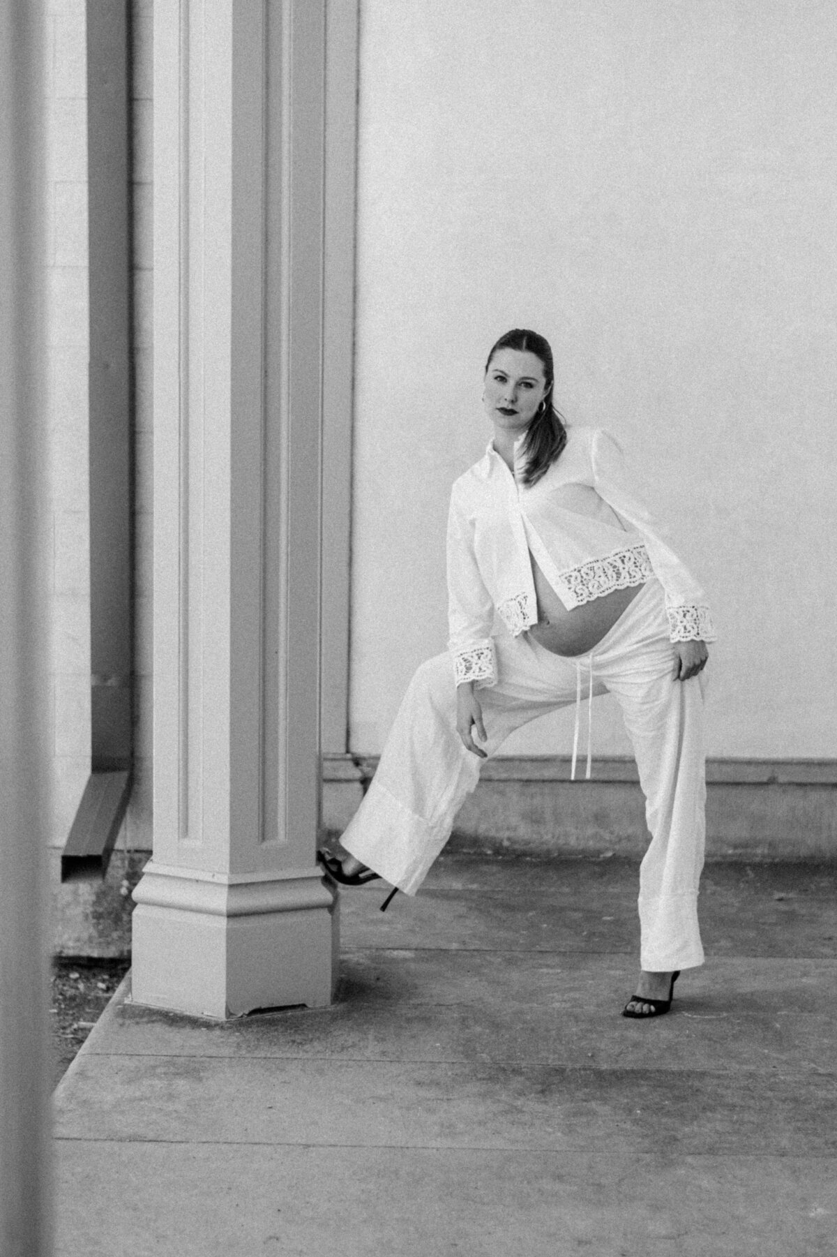 Editorial black-and-white maternity portrait of a poised mother-to-be in heels, framed by architectural columns