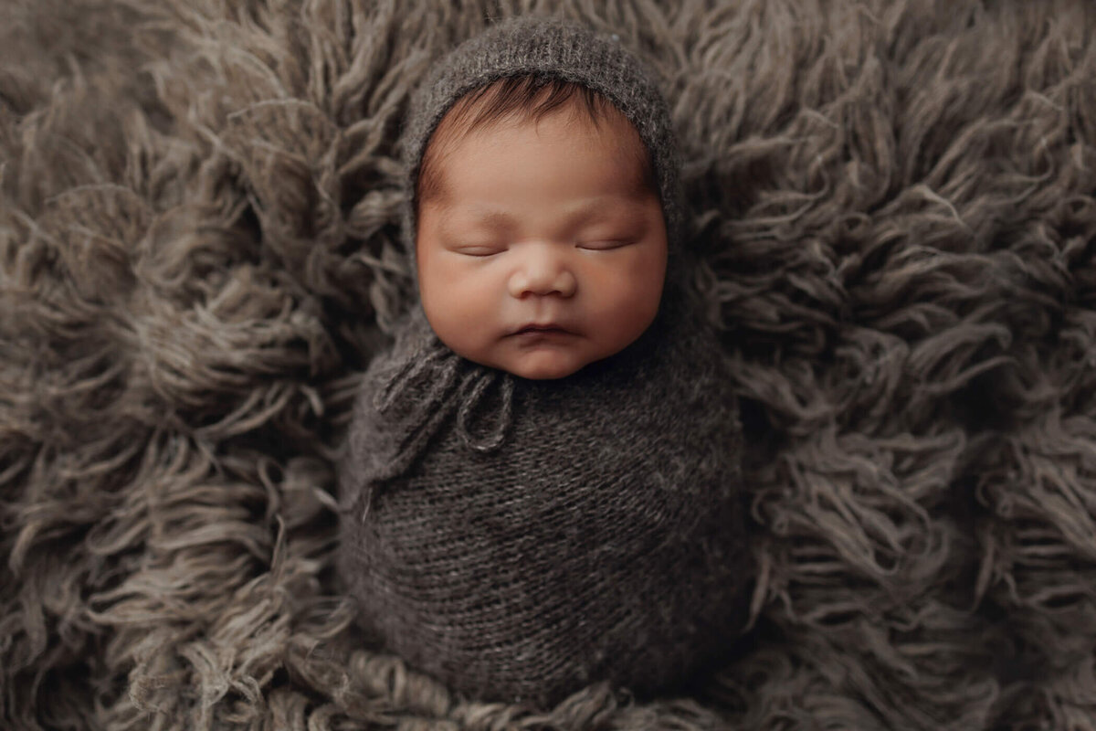 A newborn baby, peacefully sleeping, is swaddled in a cozy, dark gray wrap with a matching bonnet. The baby is nestled on a fluffy, textured gray blanket, creating a soft and warm atmosphere.