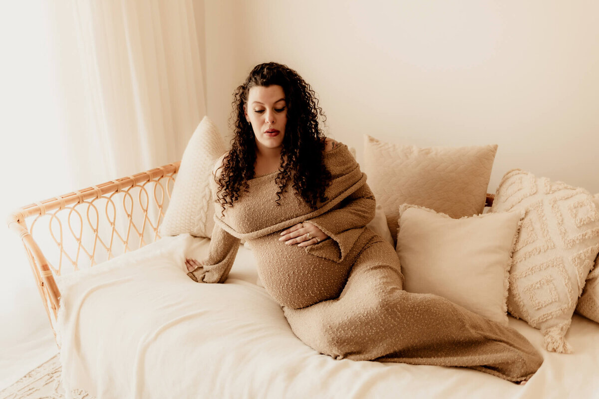 Pregnant woman in OKC wearing a beige sweater dress and looking down at her belly on a daybed.