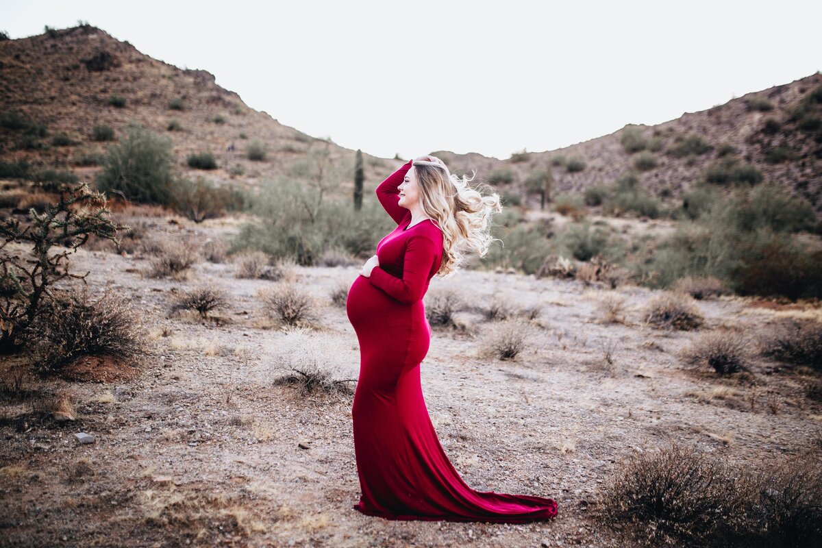 maternity photos with woman in a red dress while holding her bump and touching her hair in a desert