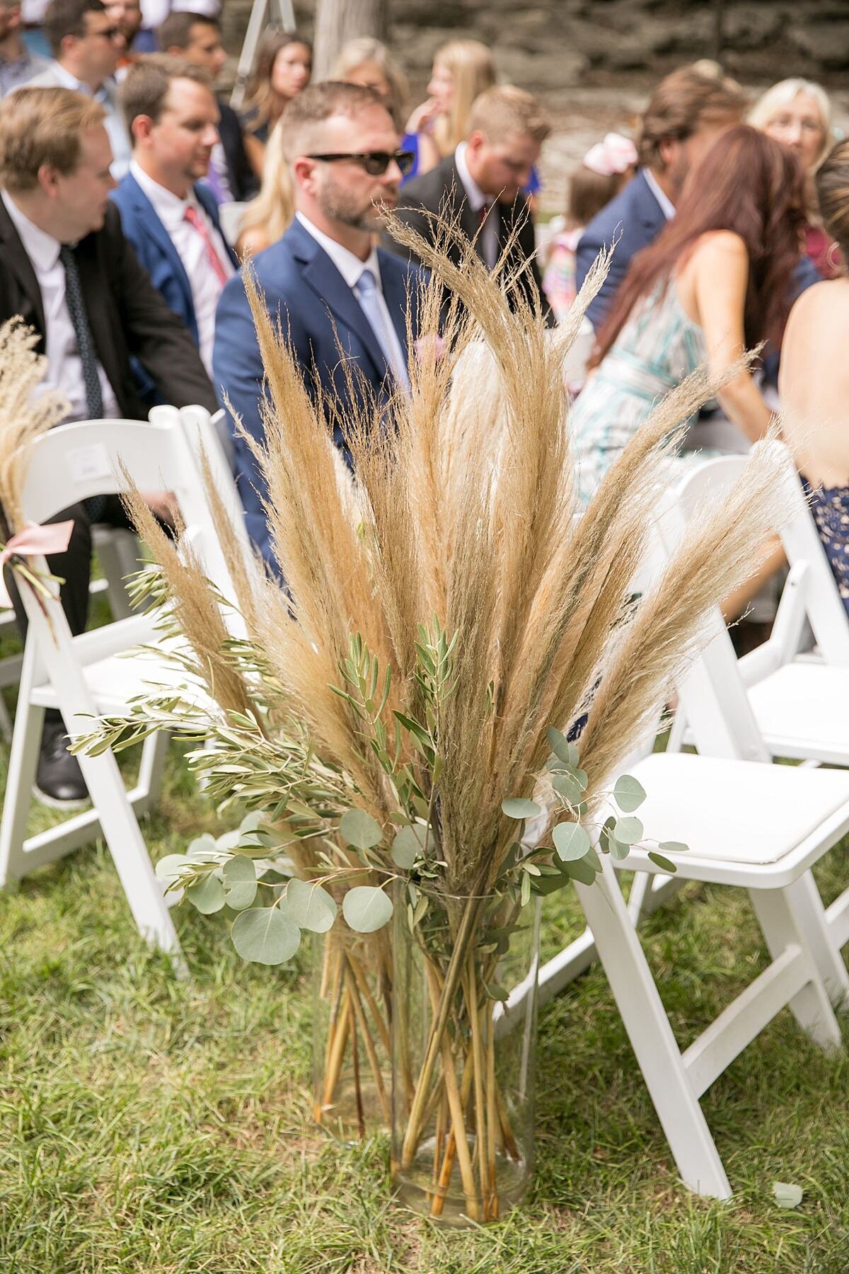 White garden chairs are decorated with large plumes of pampas grass, greenery and eucalyptus as they decorate the aisle for this boho wedding at Saddle Woods Farm
