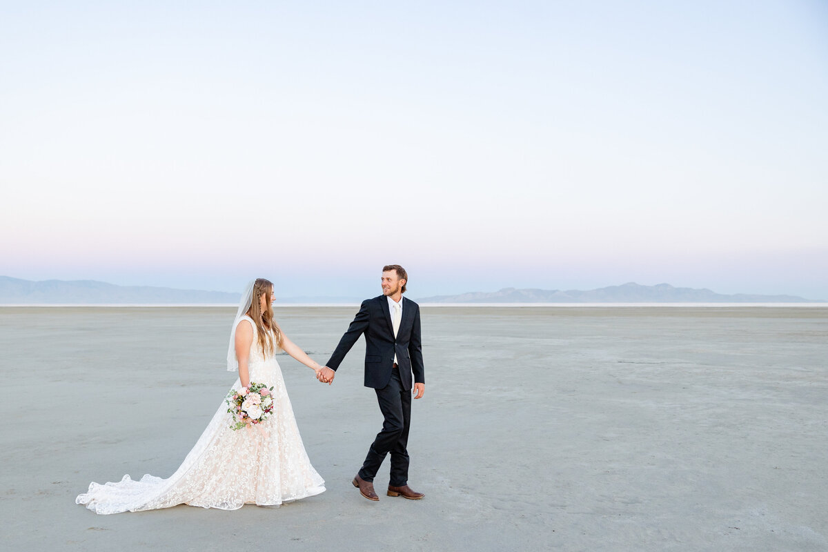 Bride and groom walk hand in hand on the Great Salt Lake’s dry flats, with distant mountains in the background.