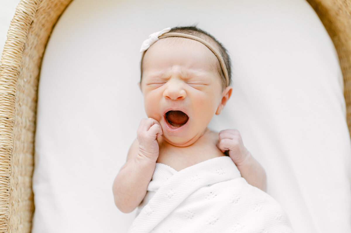 Baby in a moses basket, yawning, with a white swaddle wrapped loosely around her chest