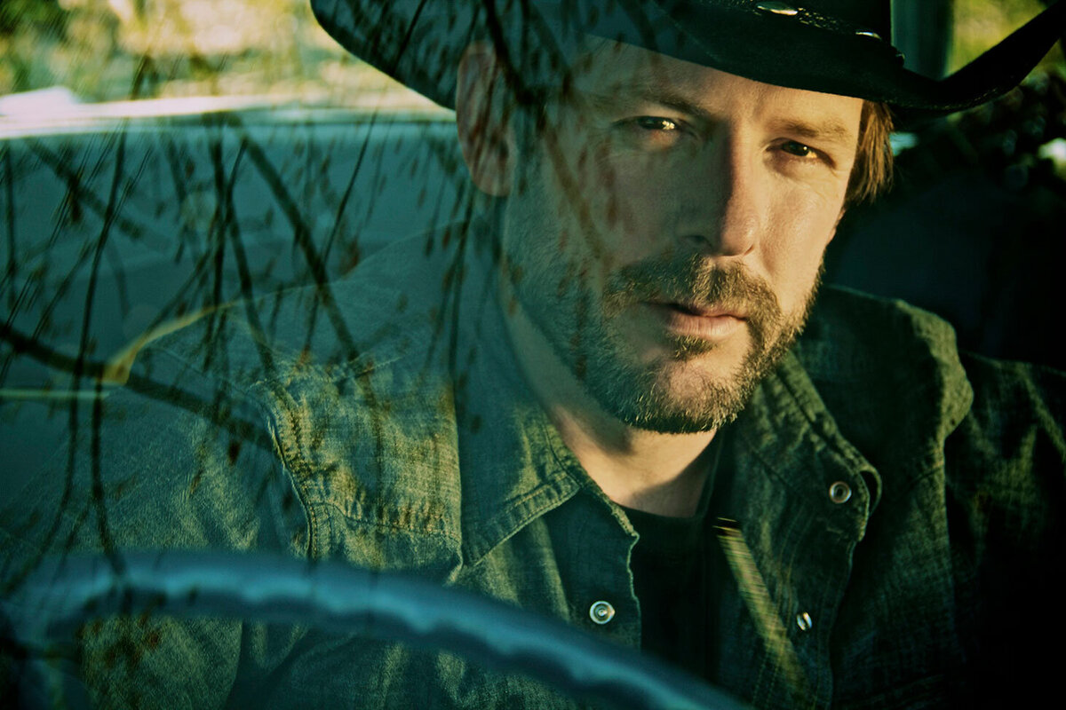 Country Musician portrait Jason Price sitting behind steering wheel of car wearing jean jacket and black cowboy hat reflection of trees on windshield Reflections Gallery