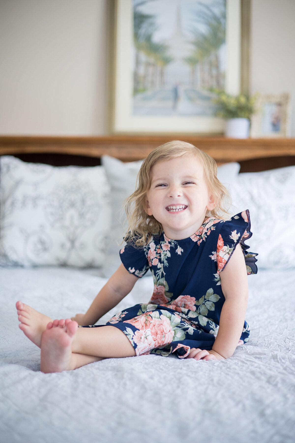 Happy toddler girl in a blue flowered dress smiling on a bed during her session with las vegas miletsone photographer