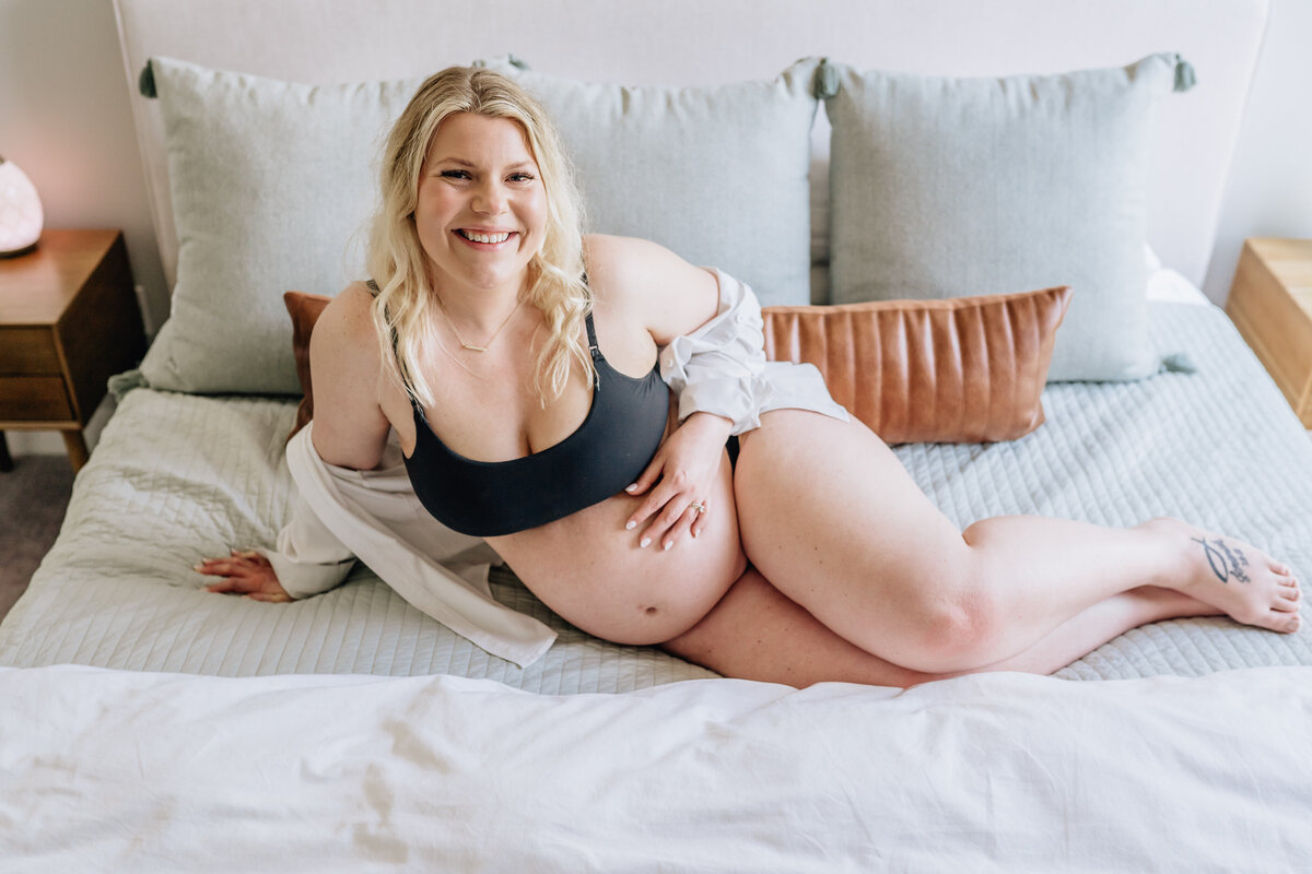 A pregnant woman reclines on a bed with light-colored pillows, smiling brightly at the camera while dressed in a black bra and white shirt.