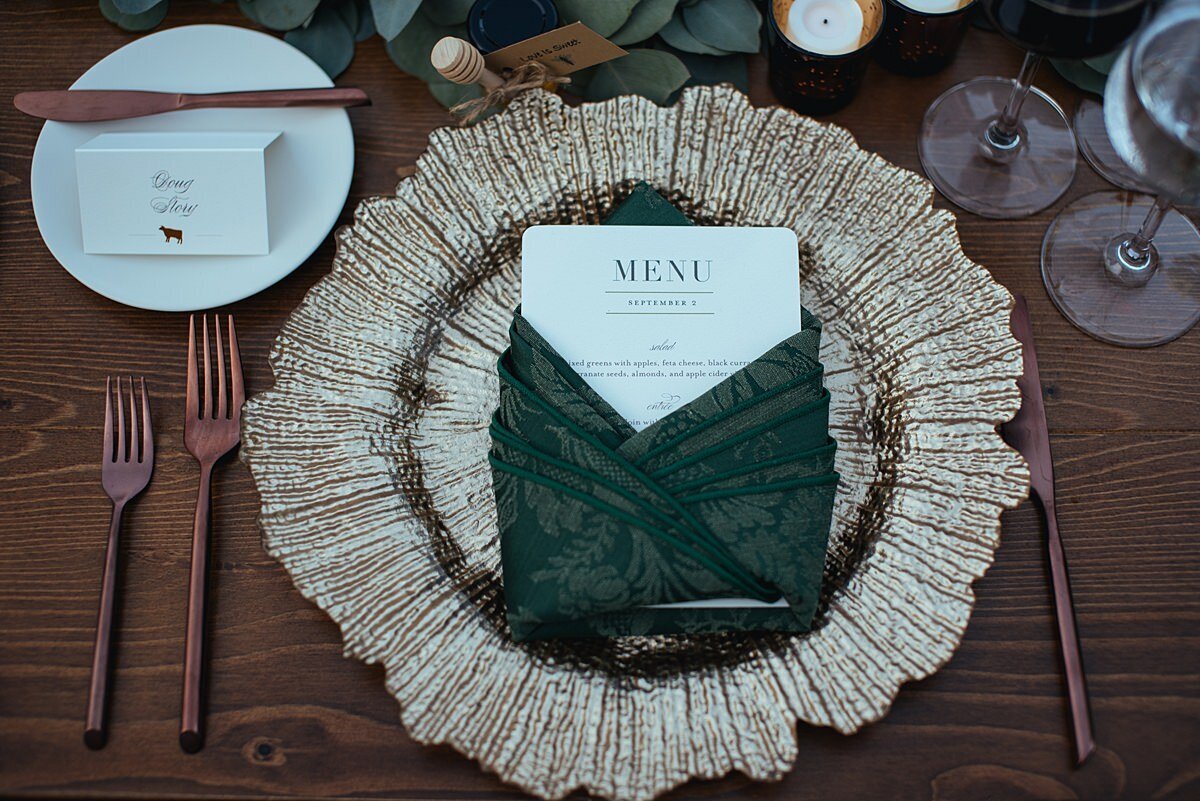 An ivory menu card is wrapped in a forest green brocade linen napkin placed on top of a glass charger that looks like a flower. Next to the charger is an ivory bread and butter plate with a guest seating card and small matte gold bread and butter knife and two stemmed wine glasses with ice water and red wine.