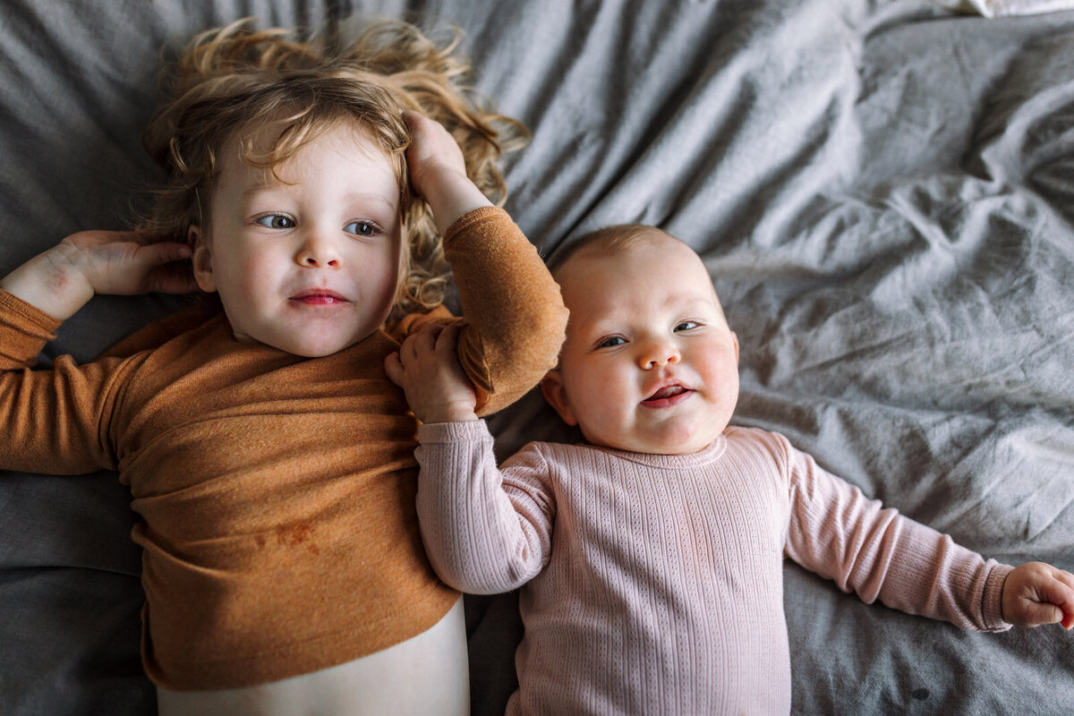 birds-eye view of toddler boy and baby sister laying on bed  on their backs with baby's arm on his arm