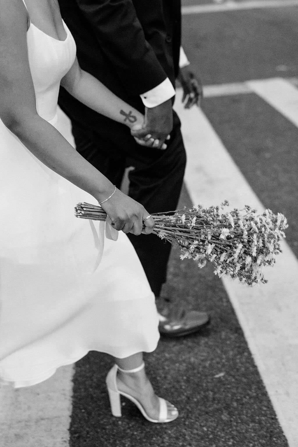 black and white photo of a bouquet of flowers behind held by a bride as she holds hands with her groom and walks across a street