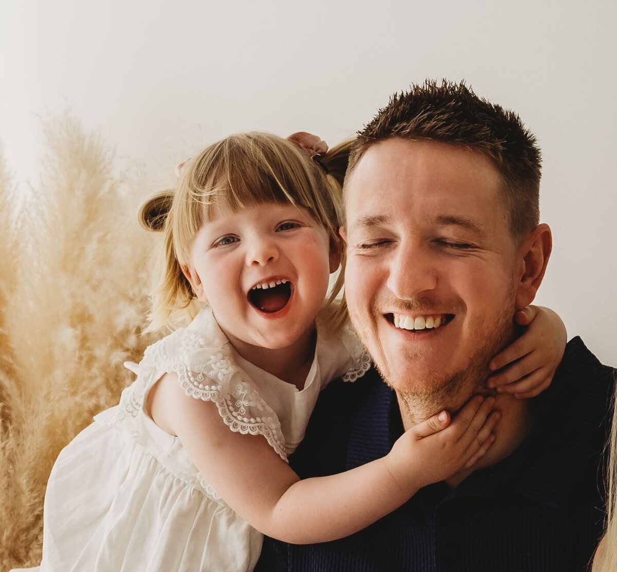 A joyful toddler girl hugs her laughing father from behind in a warm studio portrait by Ruaa Hamdan Photography