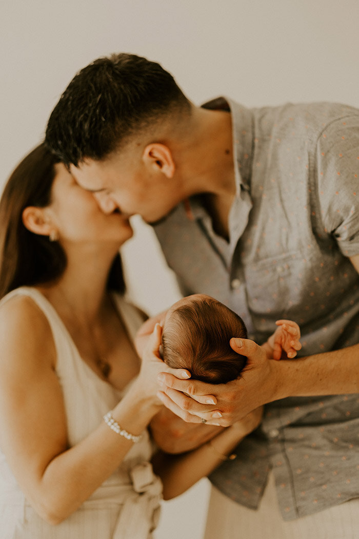 Couple échangeant un baiser et tenant dans leurs mains leur bébé au premier plan lors d'une séance photo naissance.