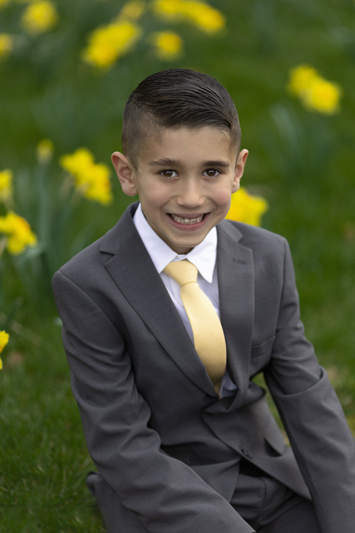 A young boy sits in a lawn with wild daffodils in a grey suit and gold tie