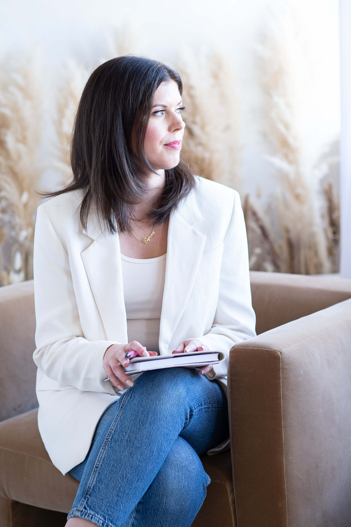 a woman in a white blazer sitting in a chair with a journal in her lap looking pensively out the window.  Captured at Skye Studio by Ottawa branding photographer JEMMAN Photography