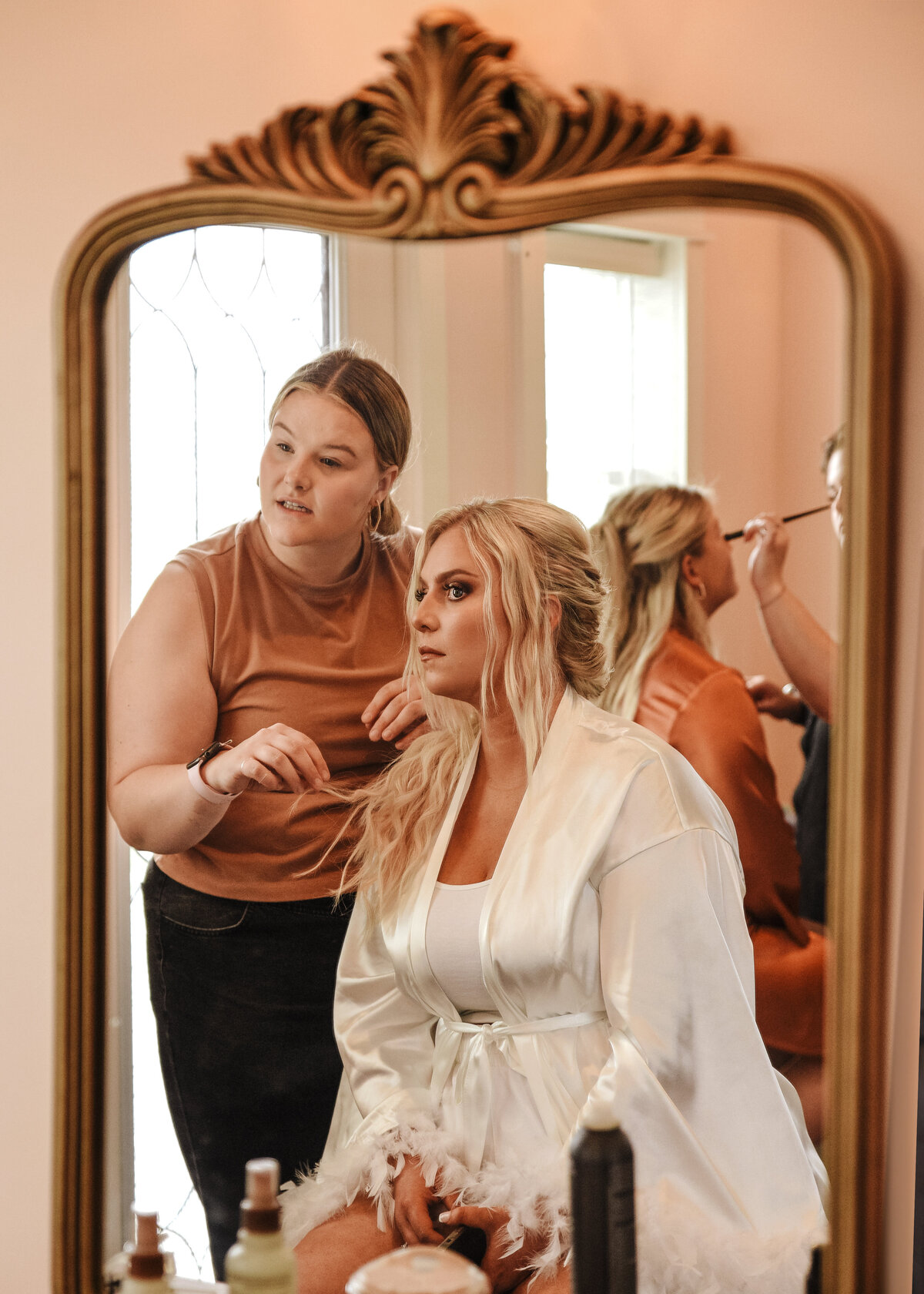 A makeup artist attentively applies cosmetics to a woman seated in front of an ornate mirror, as they both focus on perfecting the look for a special occasion taken by jen Jarmuzek photography a Minneapolis wedding photographer