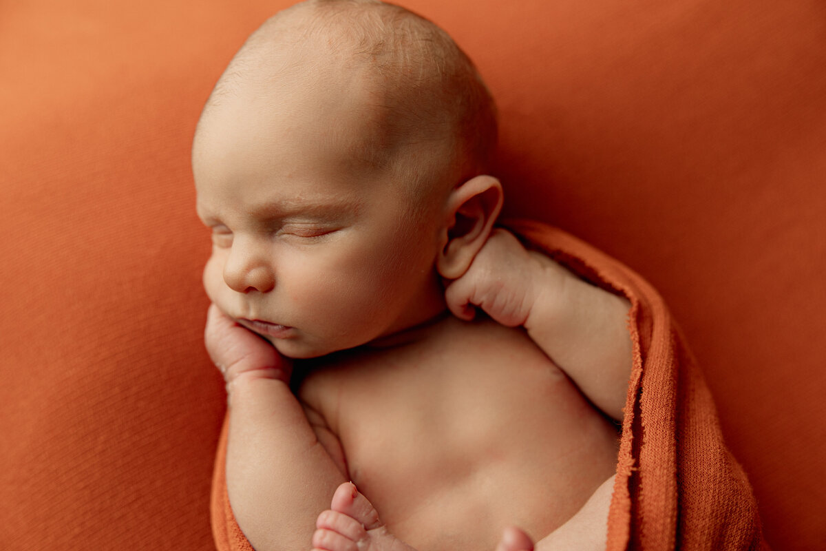 Sleeping baby boy wrapped in rust-colored fabric on a matching backdrop, highlighting warmth and autumn tones. Captured in-studio by Inland Empire newborn photographer Kaitlyn Dawn.