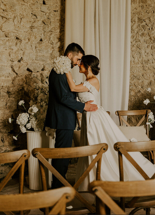 Mariés front à front posant lors d'un shotting photo mariage dans leur salle de cérémonie en vieilles pierres décorée de chaises en bois et de voilage .