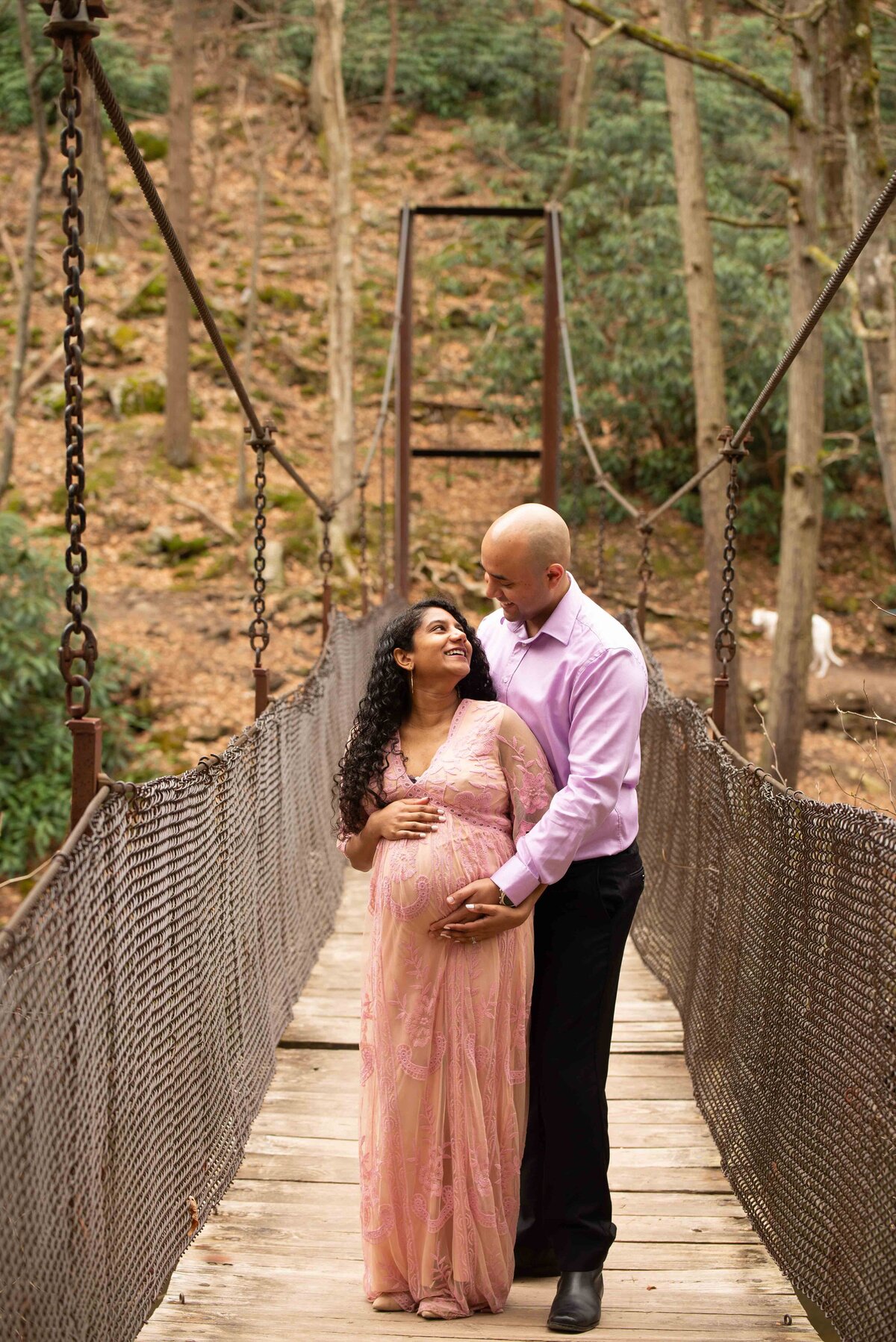 A couple stands on a bridge, with the man standing behind a pregnant woman, holding her baby bump