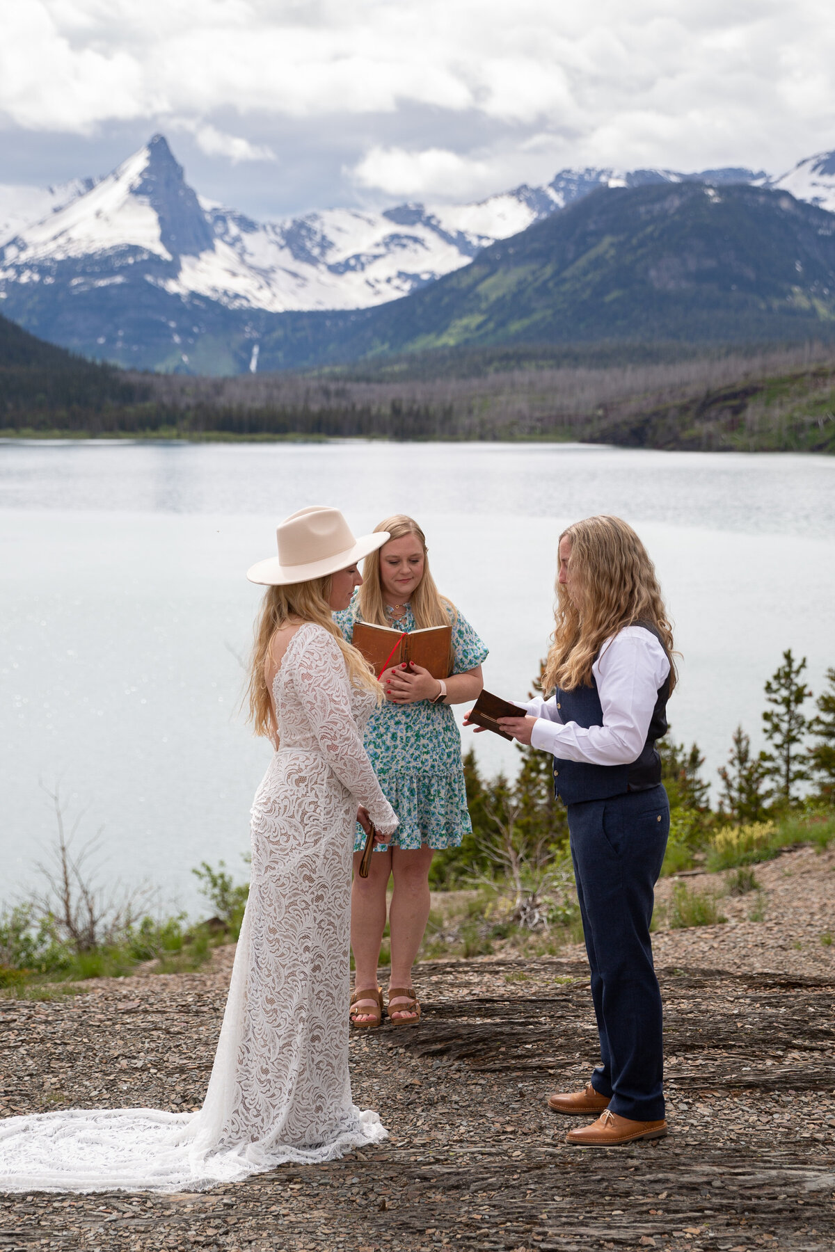 Two brides and their officiant stand together on top of a dirt path with a big lake and snow capped mountains behind them.