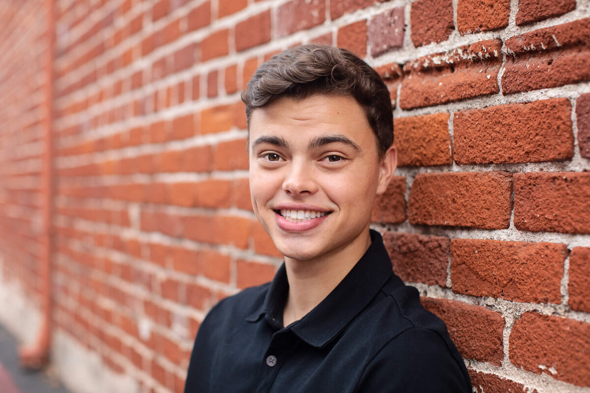 A smiling person with short brown hair leans against a brick wall. They are wearing a black polo shirt, and the background features a red brick pattern.