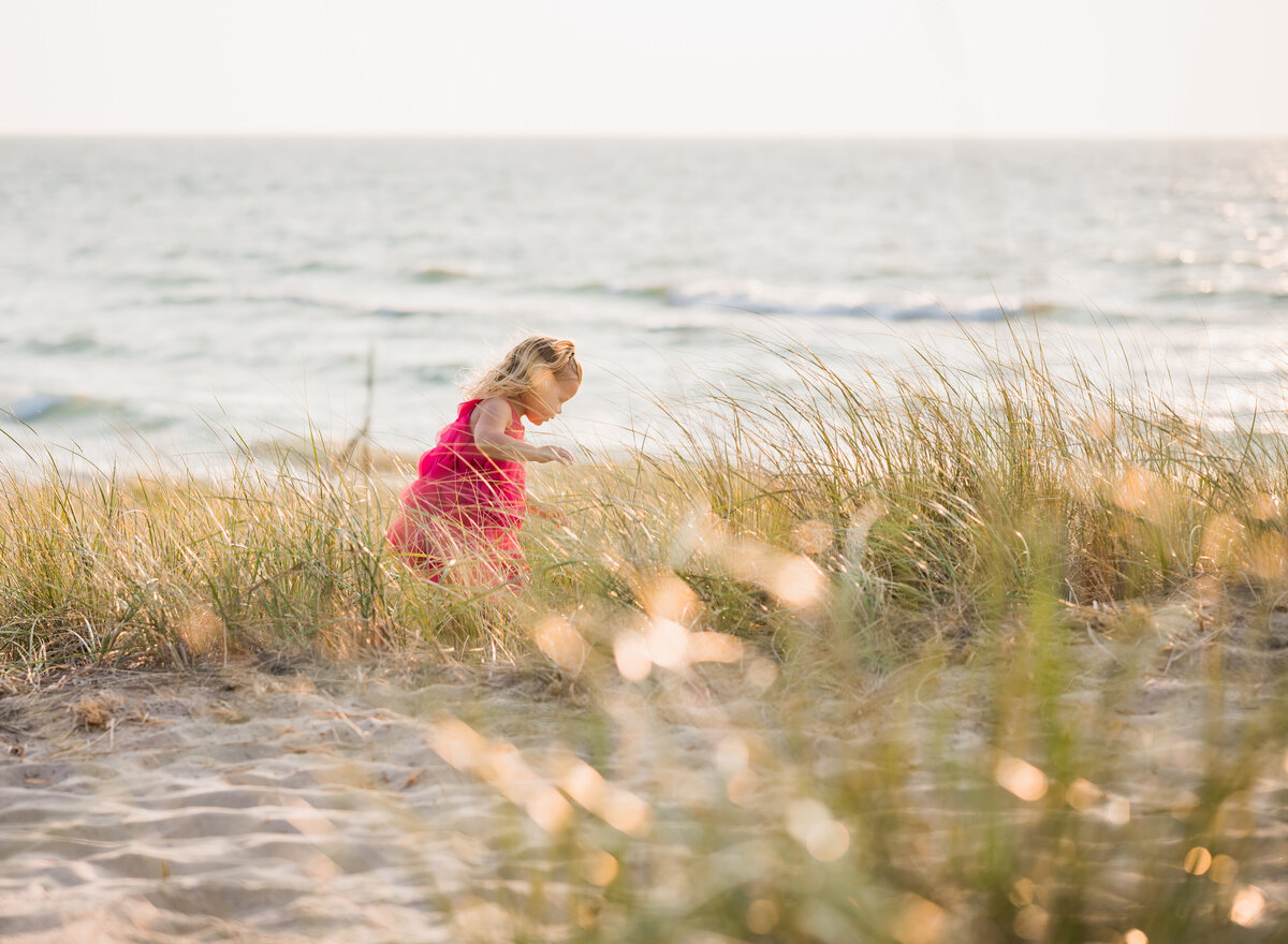 "A young girl in a pink dress walking along the beach, surrounded by tall grass and the ocean in the background