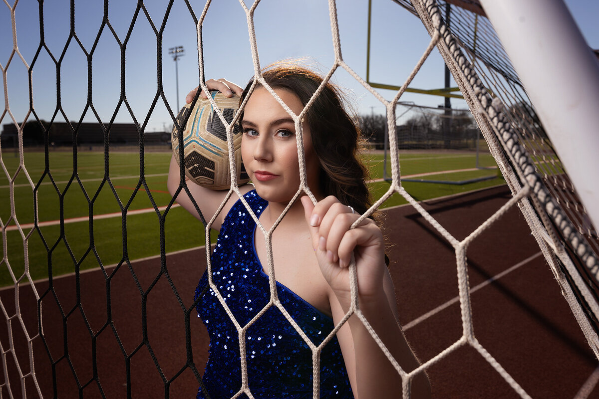 A high school senior in a blue sequin dress poses with a soccer ball for senior photography in Dallas.