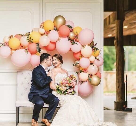 Bride and groom sitting on a white bench underneath pink, orange and gold balloon arch decorated with greenery