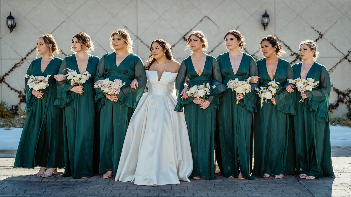 Winter wedding party at Divine Grace Vineyards in Dallas, Texas – bride in satin gown with bridesmaids in emerald green dresses holding blush and white bouquets, photographed by JPF Galleries wedding photographer.