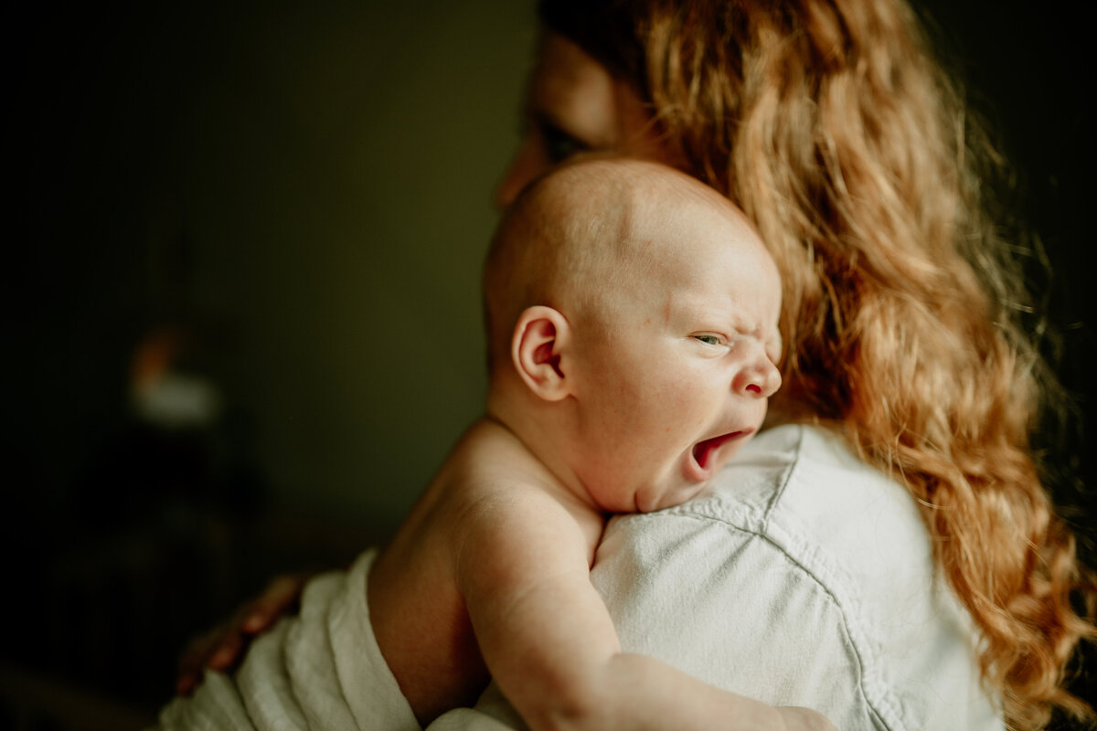 A sweet newborn baby boy yawns while perched on his mama's shoulder during an in-home lifestyle photoshoot.
