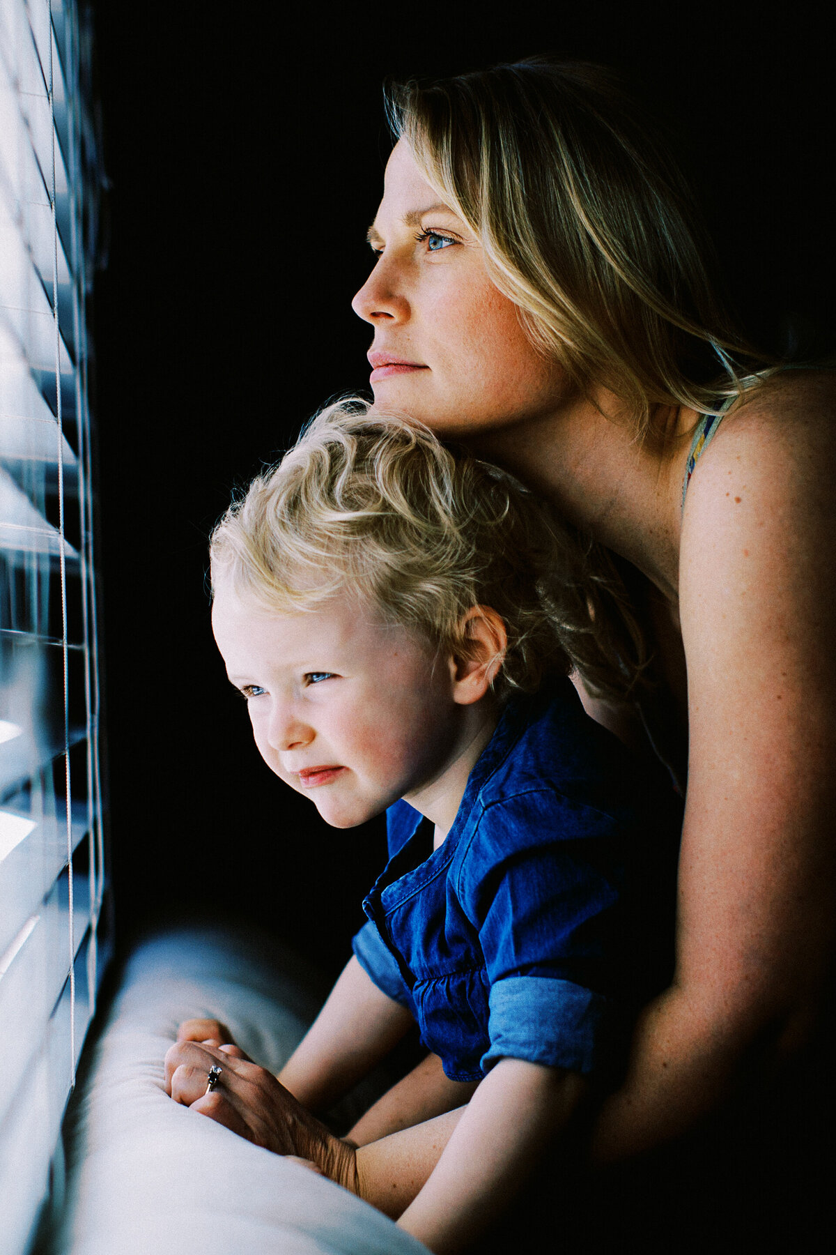 a mom rests her chin on young blonde daughter while both look out window