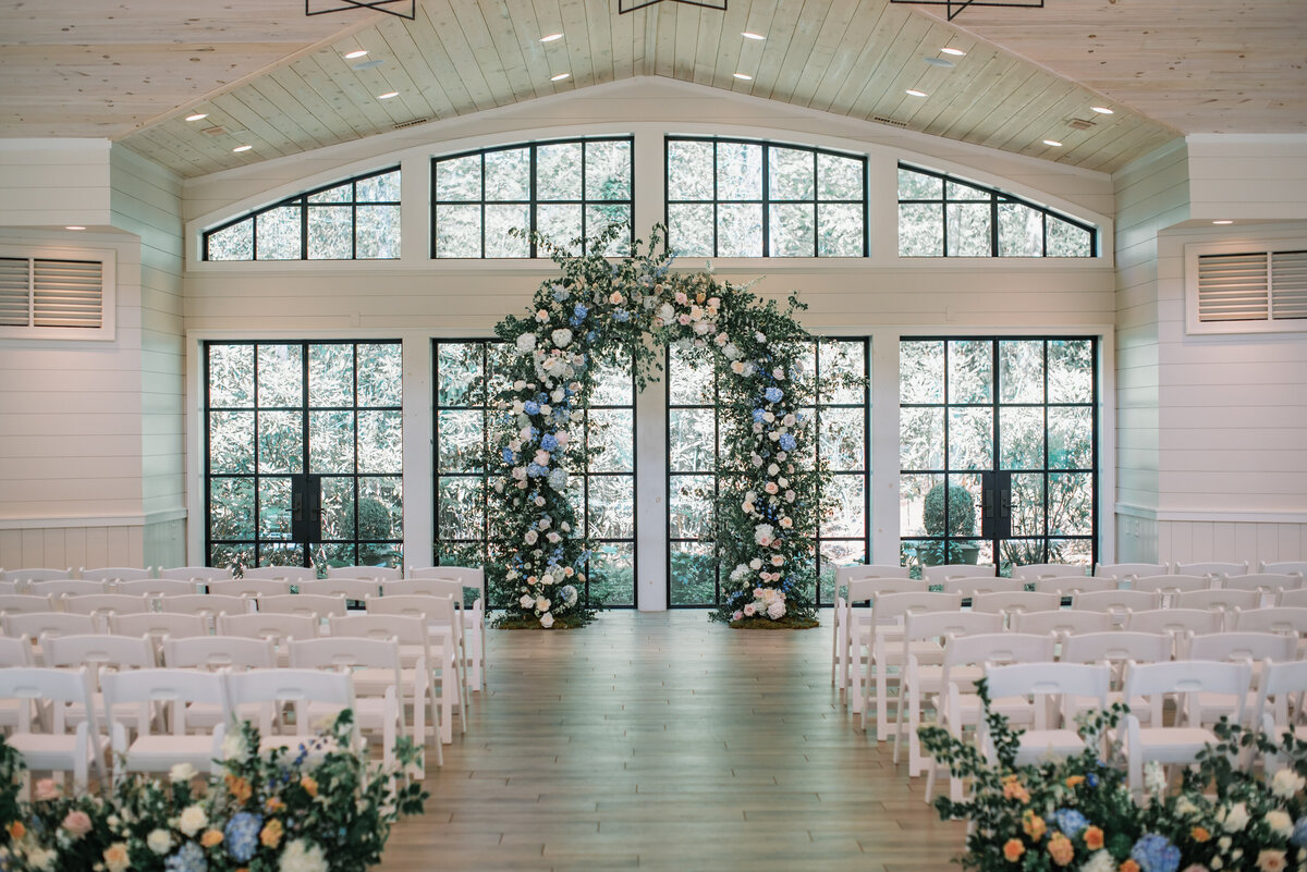Indoor wedding ceremony at Old Edwards Inn with lush floral arch in soft blue and white flowers, modern windows, and white chairs.