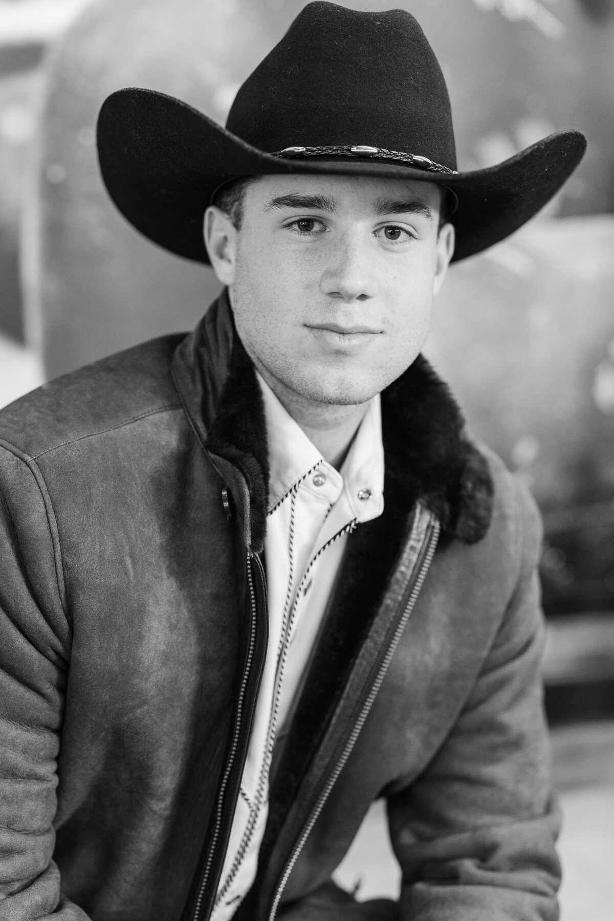 Black and white senior portrait of a young man in a cowboy hat and suede jacket, seated in Mule Alley, Downtown Fort Worth. Western-inspired style with confident, timeless expression.