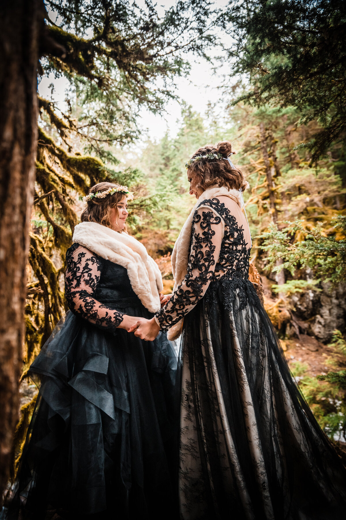 Two brides, both wearing black wedding gowns, flower crowns, and white fur stoles, hold hands during their Alaskan elopement ceremony.