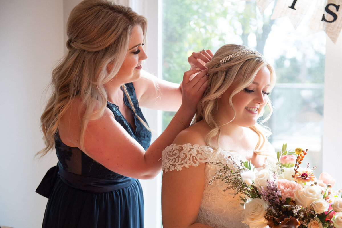 a bridesmaid in a blue dress adjusts the bride's veil while she holds a bouquet with fall colours.  Captured indoors by Ottawa wedding photographer JEMMAN Photography